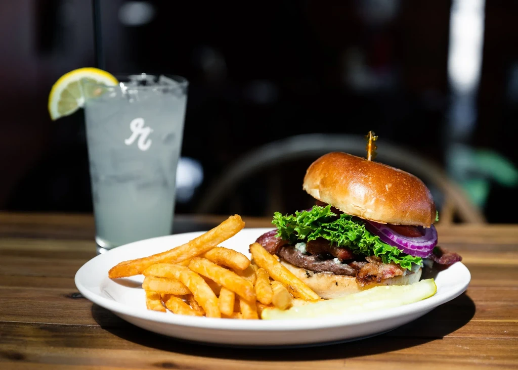 Burger, fries, pickle, and iced drink with lemon on a wooden table.