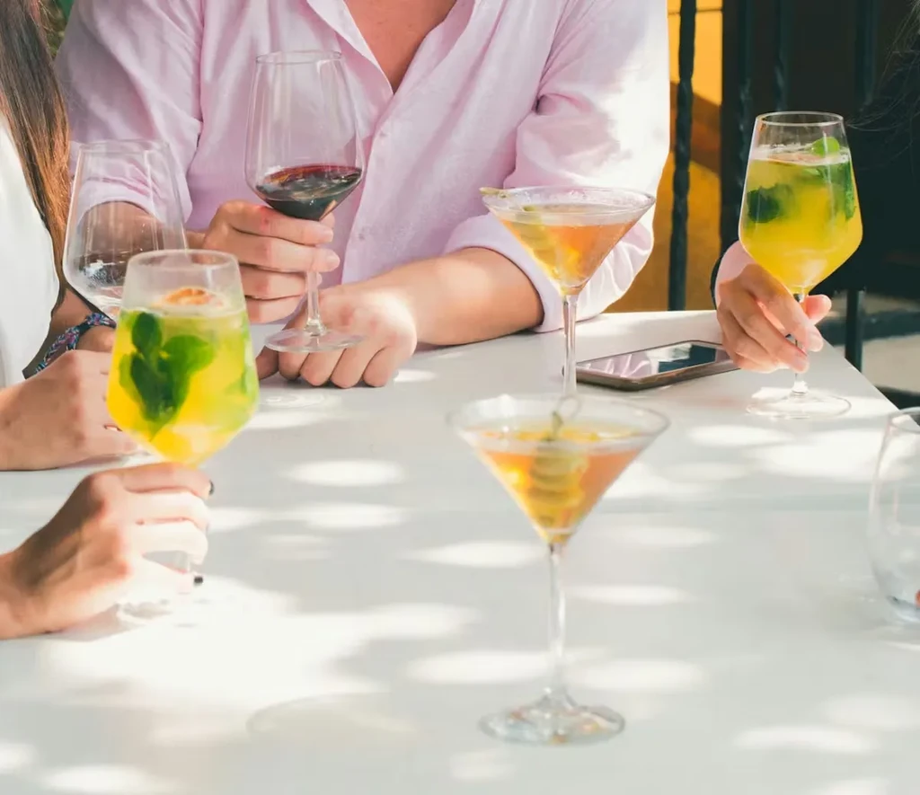 People's hands hold various cocktails and wine on a sunlit table.