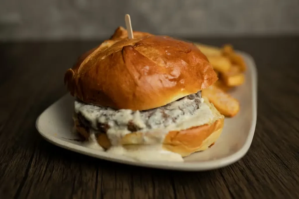 Gourmet burger with creamy white sauce, skewer, and fries on dark table.