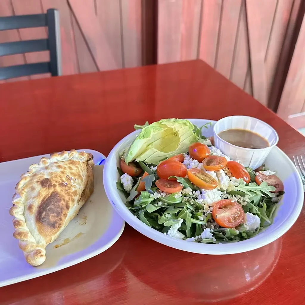 Empanada, colorful salad topped with avocado, and dressing on red table.