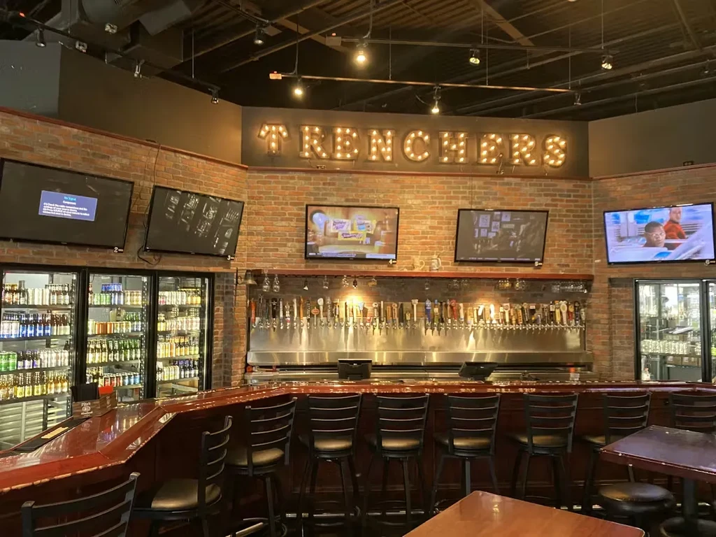 Bar interior with 'TRENCHERS' sign, long counter, and many beer taps.