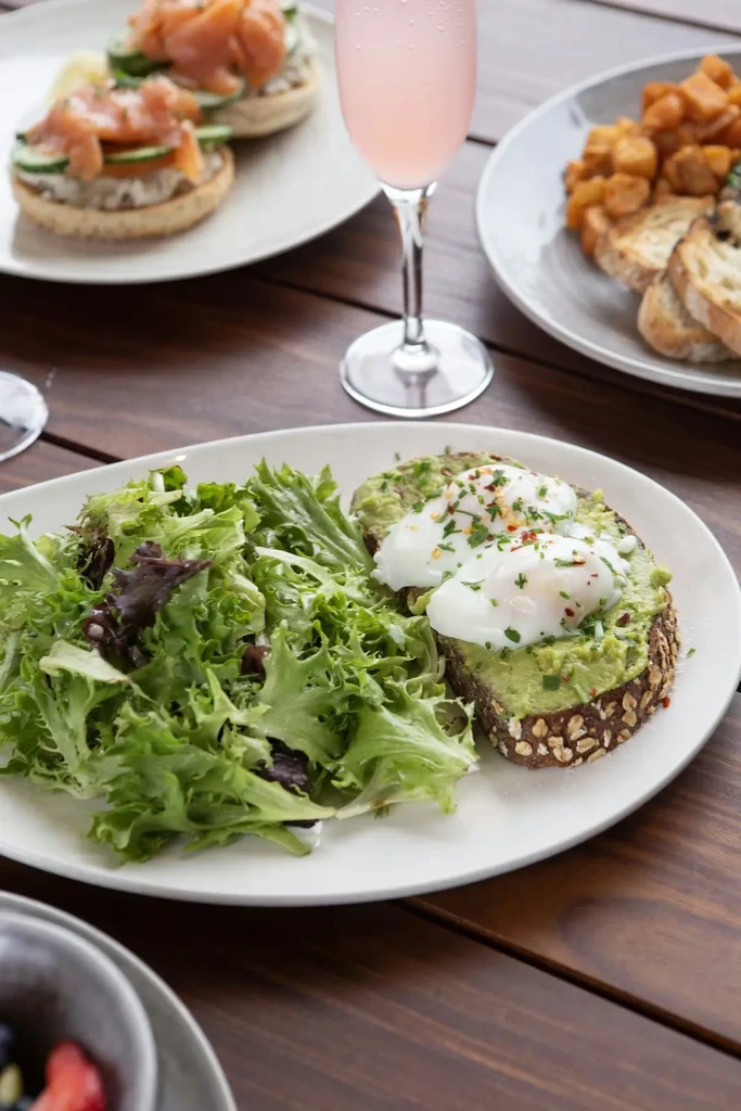 Avocado toast, poached eggs, salad, pink drink on dark wood table.