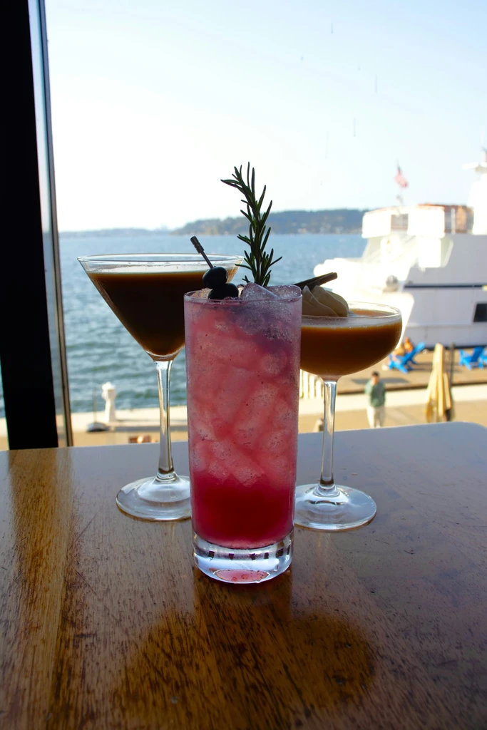 Three cocktails on a rustic table with a waterfront and yacht.