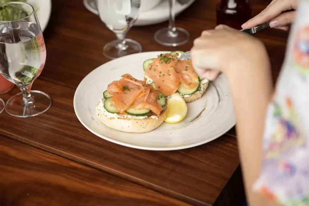 Person cutting into open-faced bagels topped with lox, cream cheese, cucumber, chives.