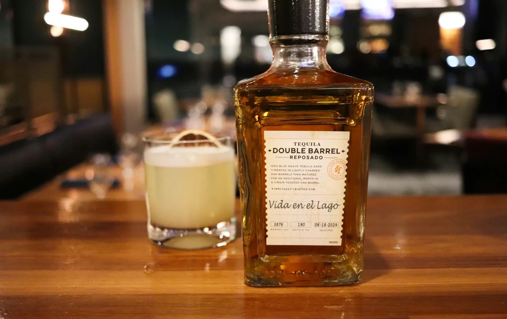 An amber reposado tequila bottle and frothy cocktail on bar.
