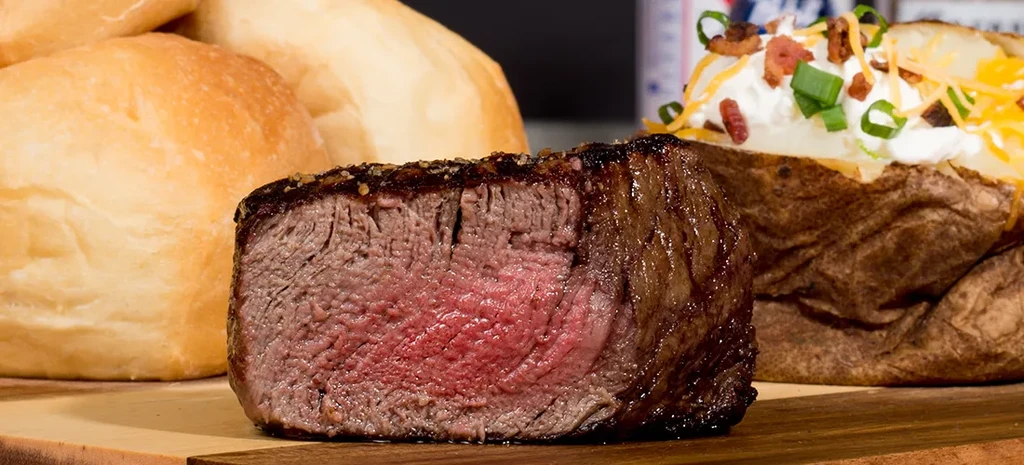 Thick cut medium-rare steak, loaded baked potato, dinner rolls on wooden board.