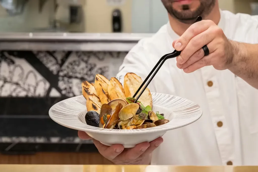 Chef holding and garnishing gourmet mussels and clams with baguette.