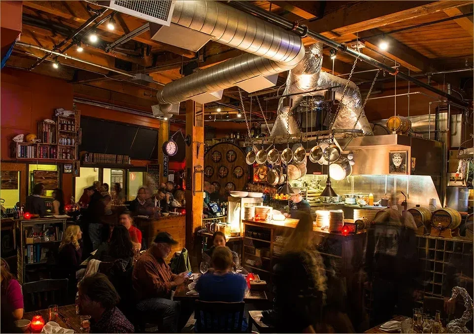 Bustling rustic-industrial restaurant with open kitchen and many diners.