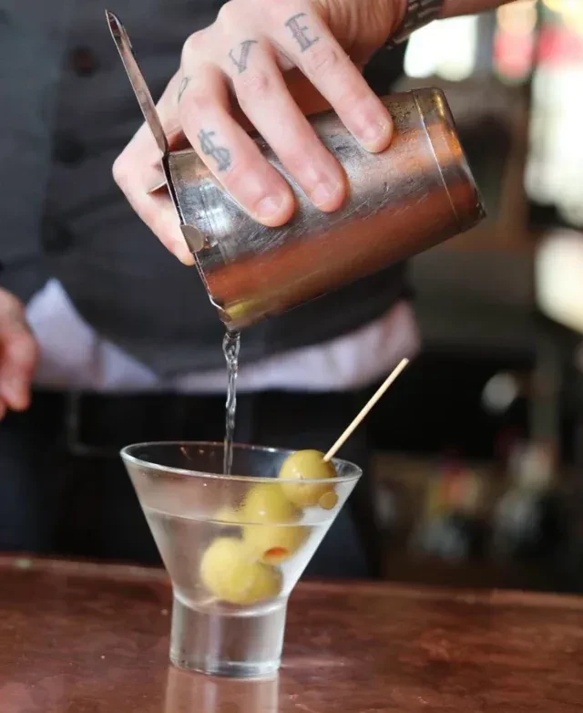 Tattooed bartender's hand pours from shaker into martini glass with olives.