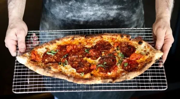 Hands hold oval pepperoni pizza with tomatoes, basil, on rack.