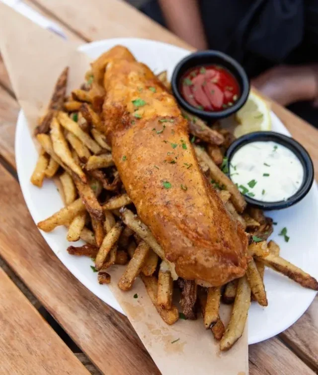 Golden-brown fish and chips, sauces, lemon, on a rustic wooden table.