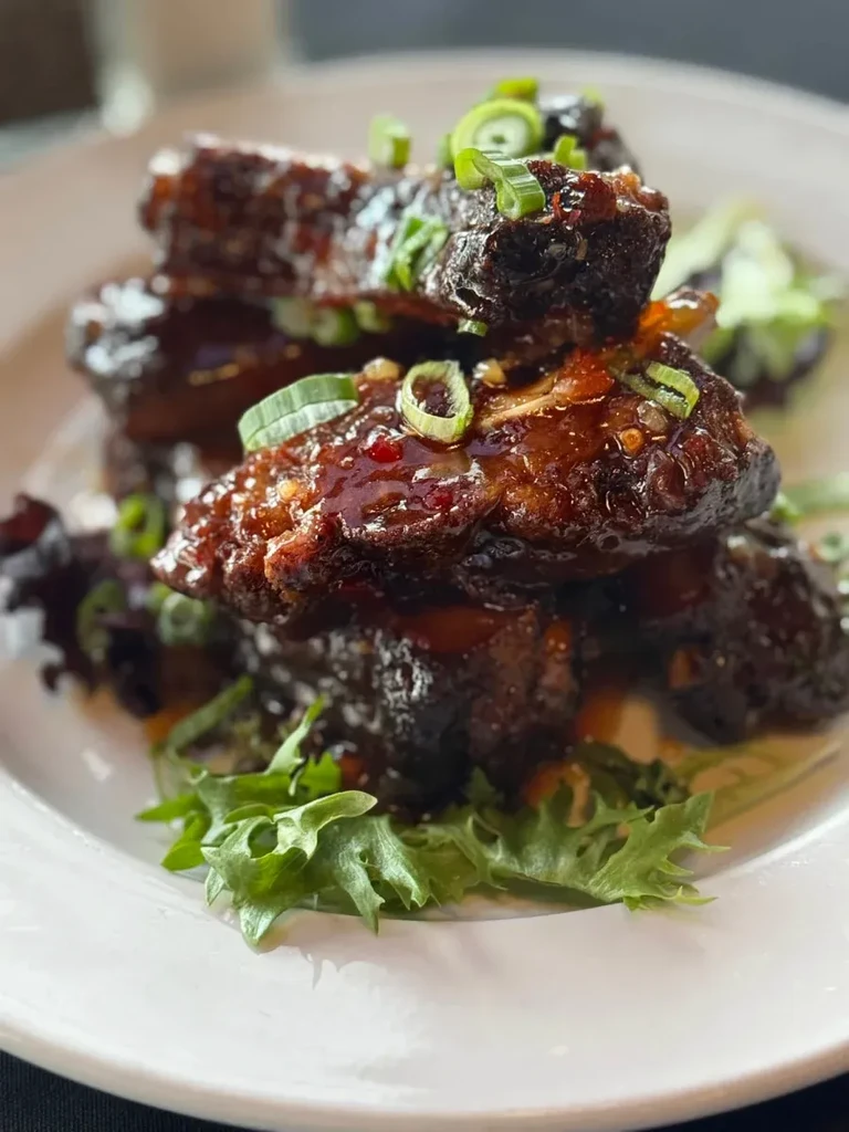 Stacked sticky glazed ribs or wings with scallions on a white plate.