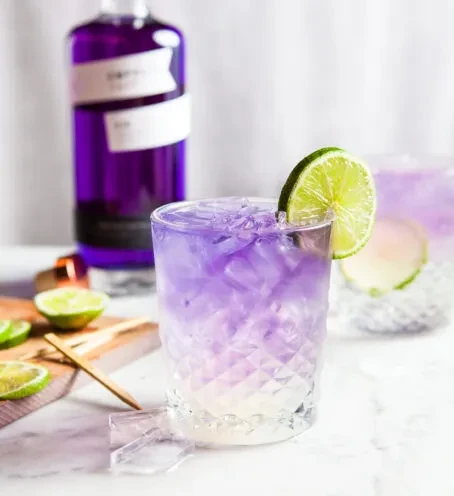 Gradient purple cocktail with lime wedge, ice, and gin bottle.