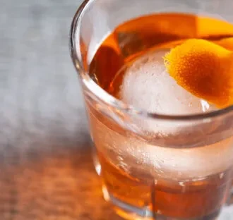 Amber cocktail in old-fashioned glass with spherical ice and orange peel.