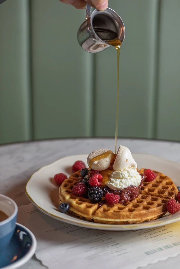 A hand pours golden syrup onto a berry-topped waffle with cream.