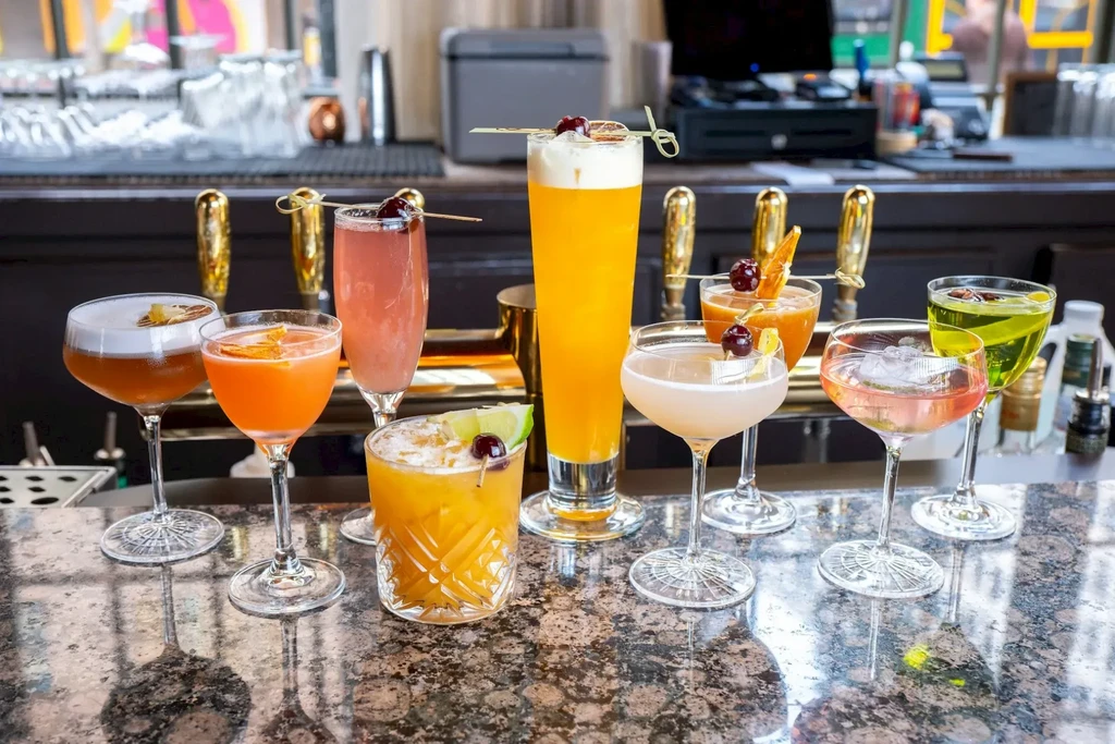 Nine colorful garnished cocktails in varied glassware on a bar.