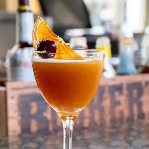 Amber cocktail in coupe glass, garnished with orange and cherry.
