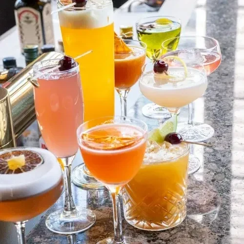 Eight colorful cocktails with garnishes in various glasses on a bar.