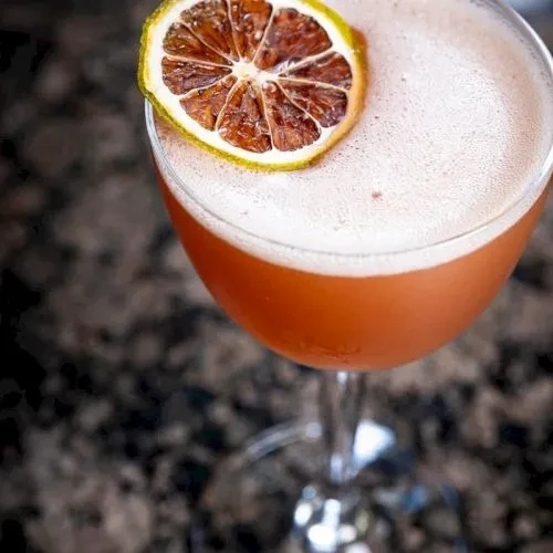 Red-orange cocktail in coupe glass with white foam and dried citrus.