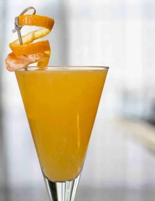 Orange-yellow cocktail in a martini glass with orange peel garnish.