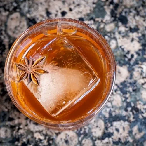 Amber cocktail with star anise and large ice in ornate glass.