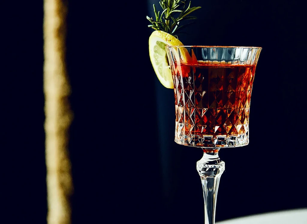 Reddish-amber cocktail in faceted crystal glass with rosemary and lemon.