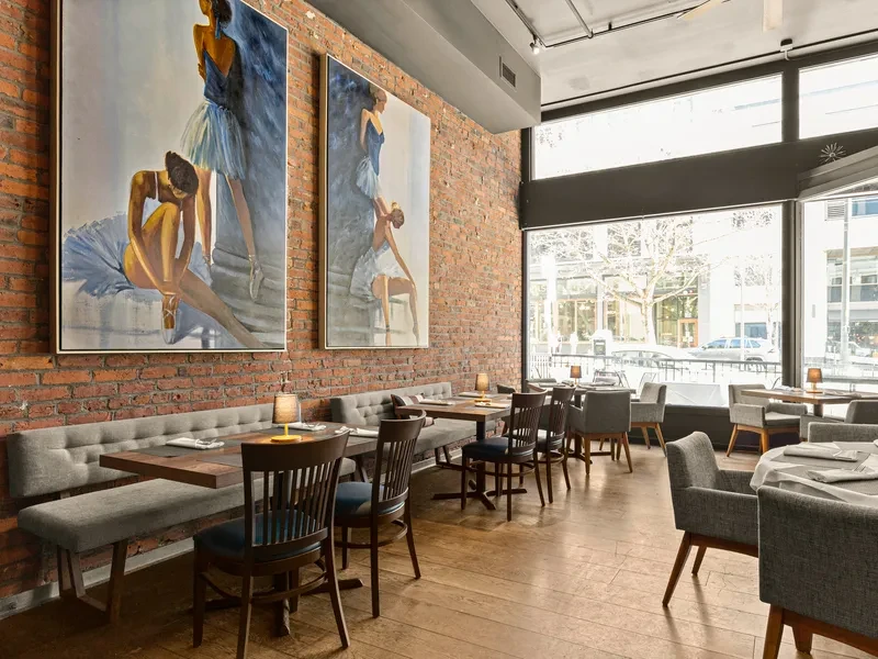 Modern restaurant with grey banquette, brick wall, ballerina art, and city view.