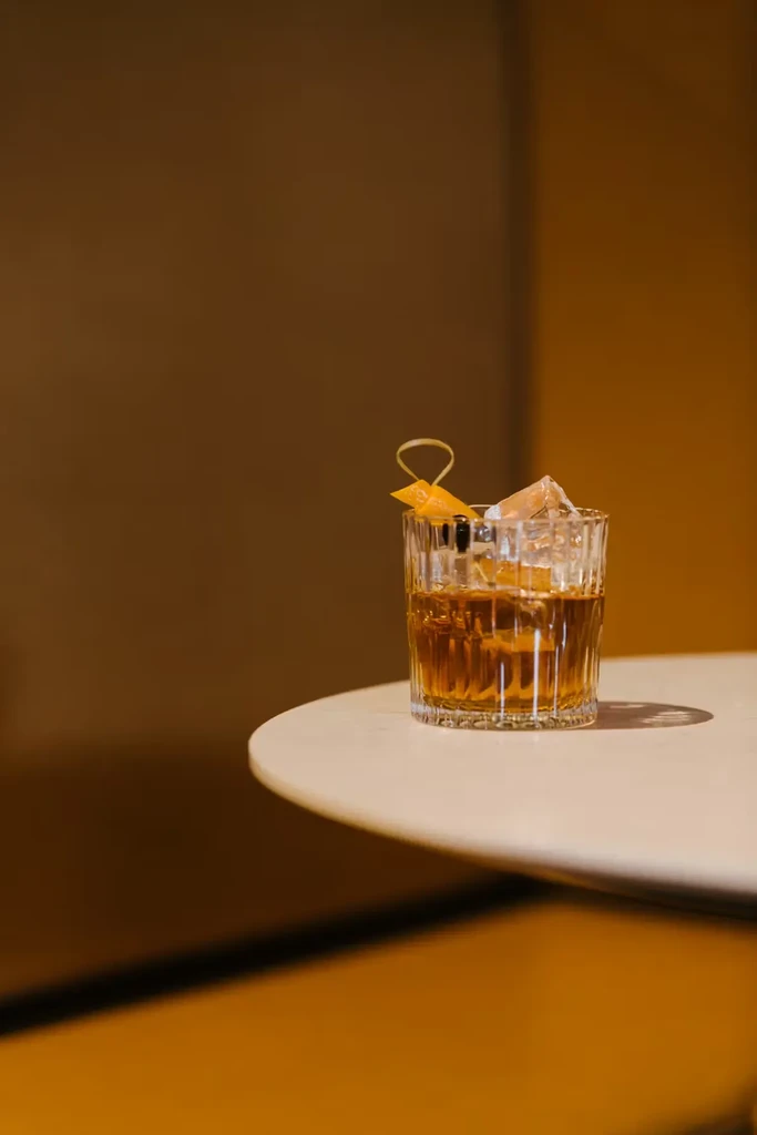 Amber cocktail in fluted glass with ice, orange peel, and cherry.