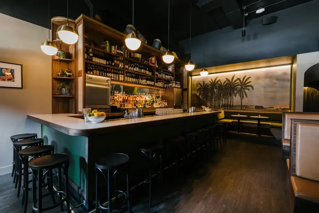 Stylish bar interior with curved counter, stools, stocked shelves, and palm tree mural.