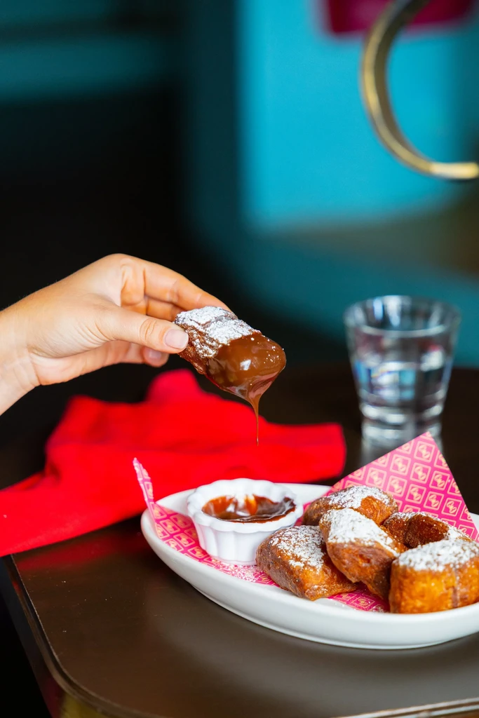 Hand dipping golden, sugar-dusted pastry into dark chocolate sauce.