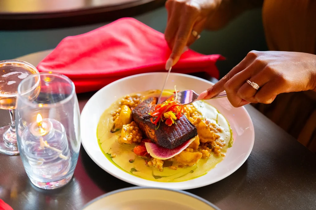 Hands cutting seared fish with grains, sauce, wine, and candle.