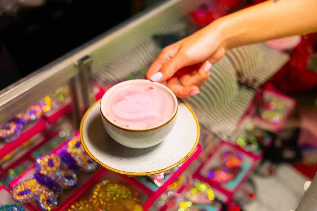 Pink-nailed hand holds grey cup with pink heart latte art; blurred background.