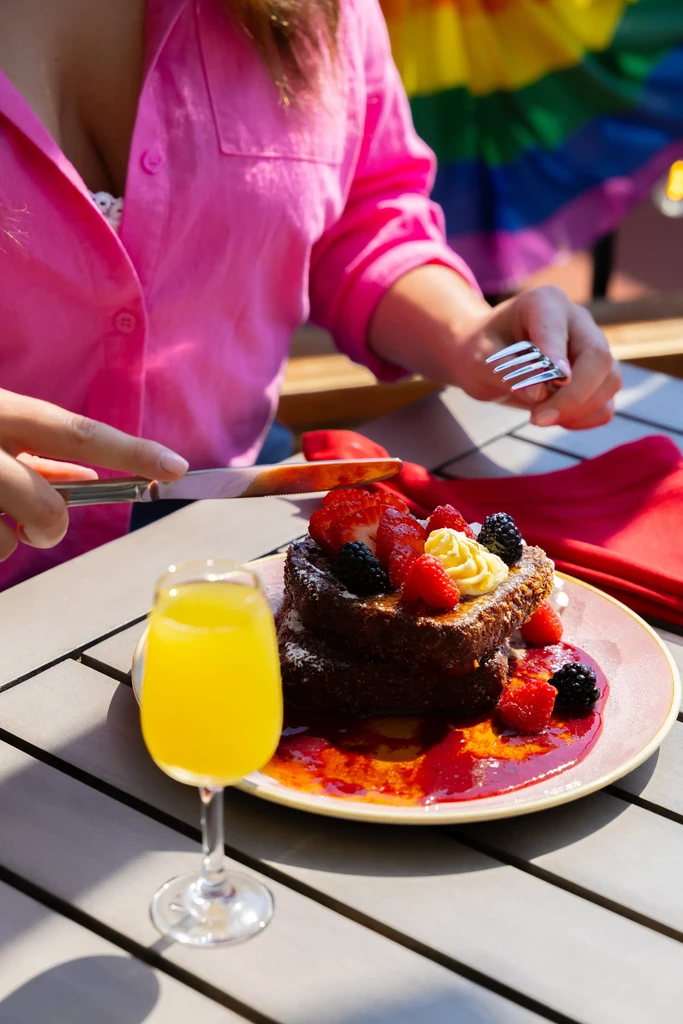 Close-up: French toast with berries and cream, mimosa, rainbow flag background.