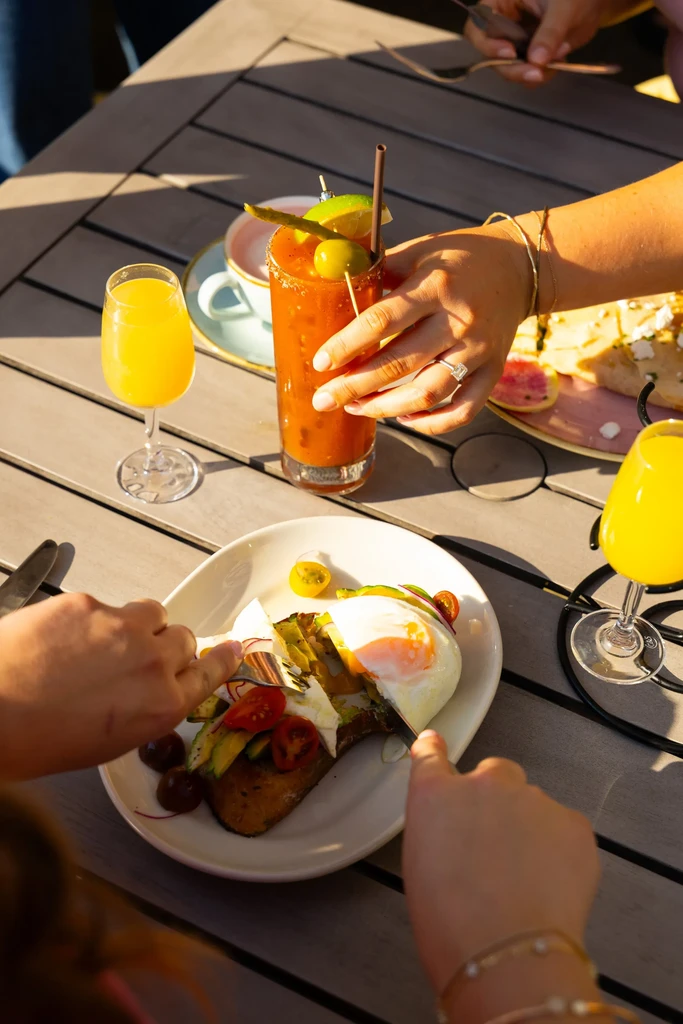 Hands eating avocado toast, mimosa, Bloody Mary at a sunny brunch.