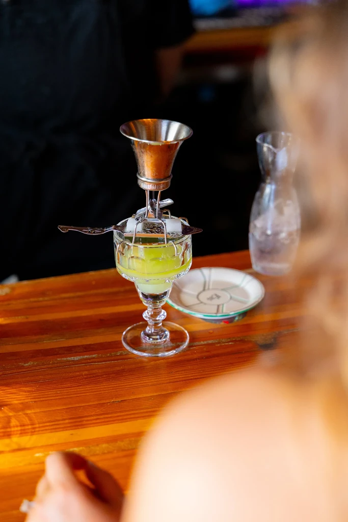 Absinthe glass, ornate spoon, sugar cubes, and water fountain on bar.