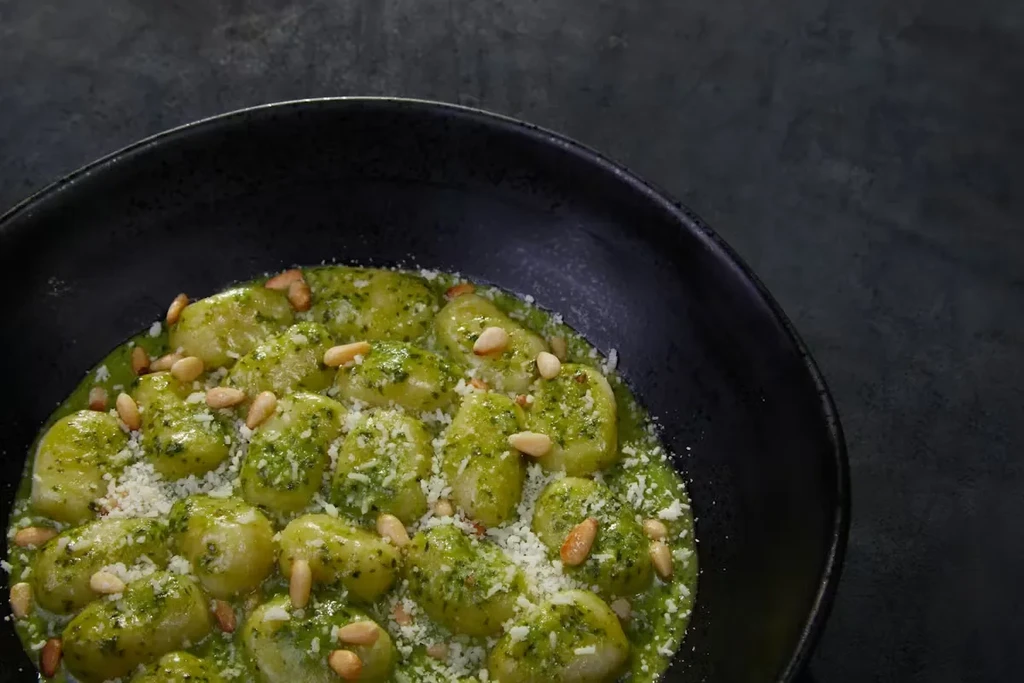 Gnocchi with green pesto, cheese, and pine nuts in a black bowl.