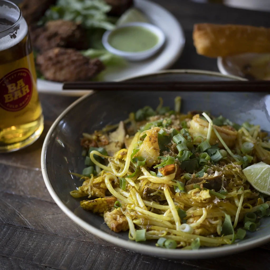 Stir-fried shrimp noodles with chopsticks, lime, beer, and appetizers on a table.