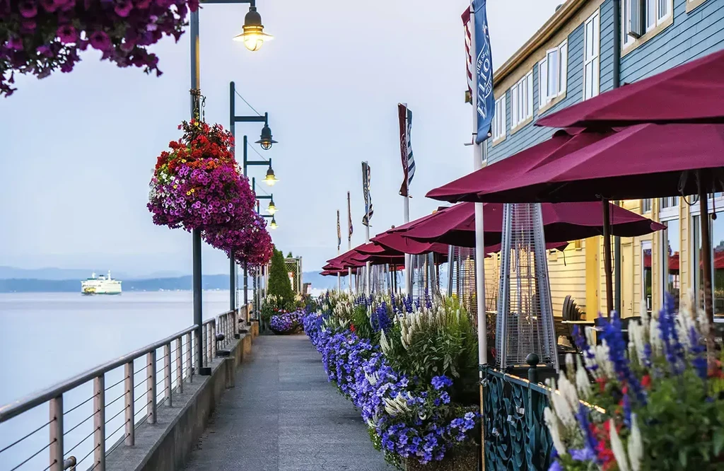 Waterfront restaurant patio with red umbrellas, flowers, and distant ferry.
