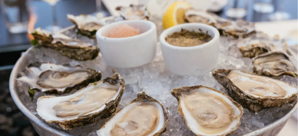 A platter of fresh oysters on ice with two sauces and lemon.