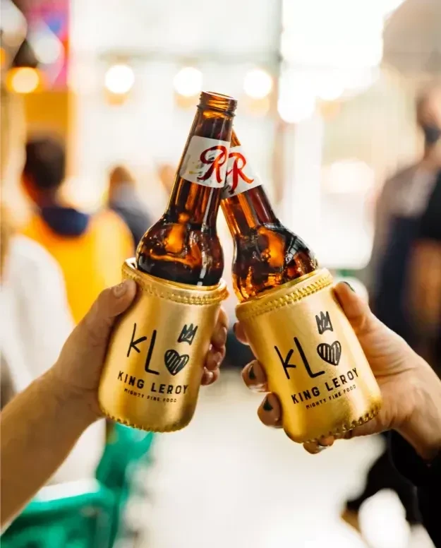 Two hands toast beer bottles in golden KL King Leroy coozies.