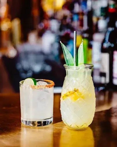 Two cocktails: one chili-rimmed, one in a pineapple glass with fruit.