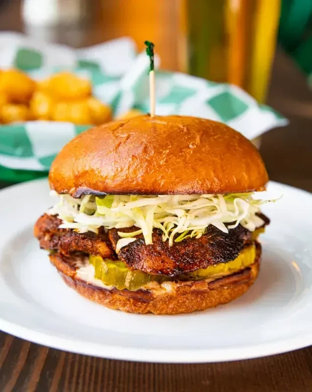 Gourmet sandwich with slaw and pickles, tater tots in background.