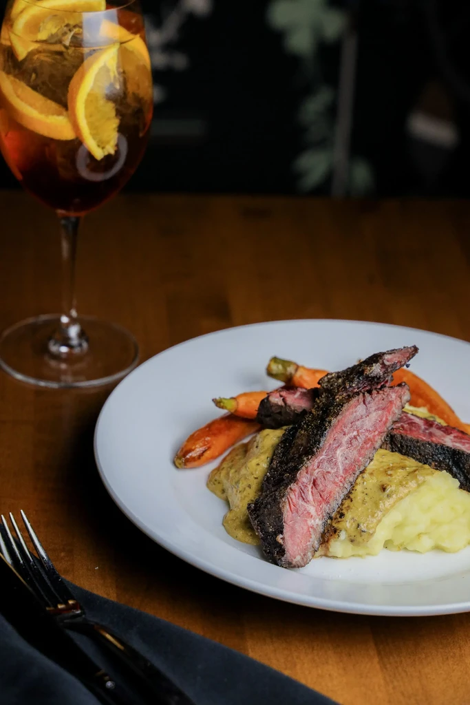 Sliced medium-rare steak with mashed potatoes, roasted carrots, and an amber cocktail.