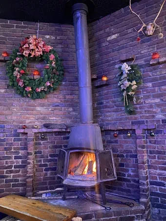 Antique wood stove with flames in a purple-lit, holiday-decorated brick corner.