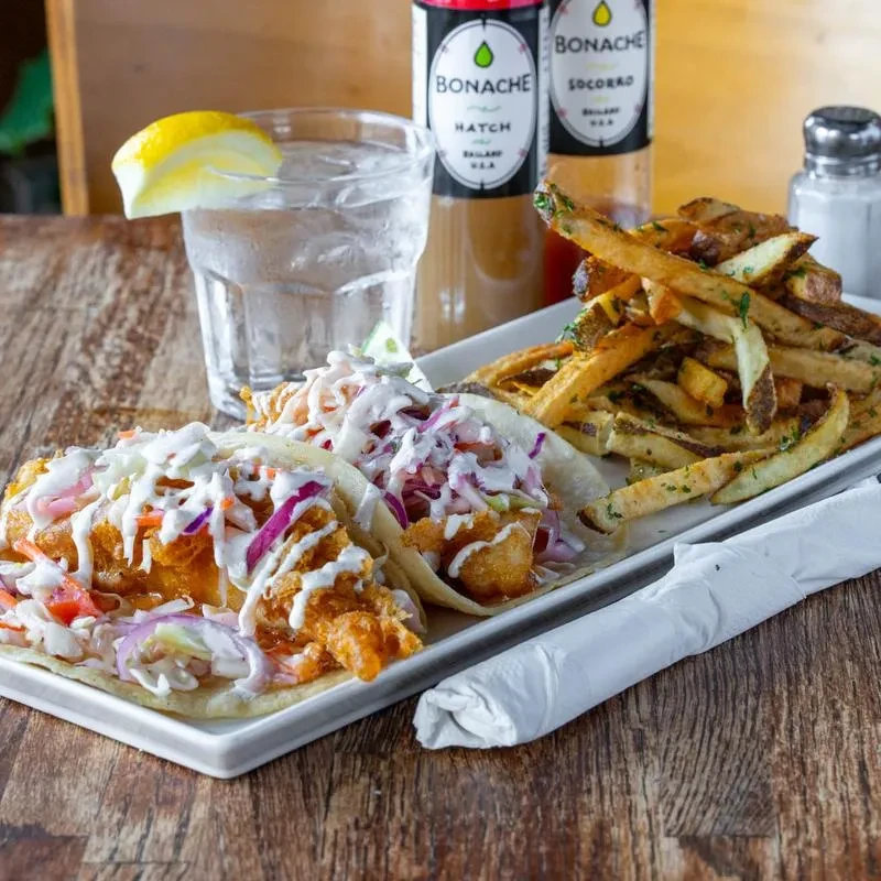 Two fish tacos with fries and a drink on wooden table.