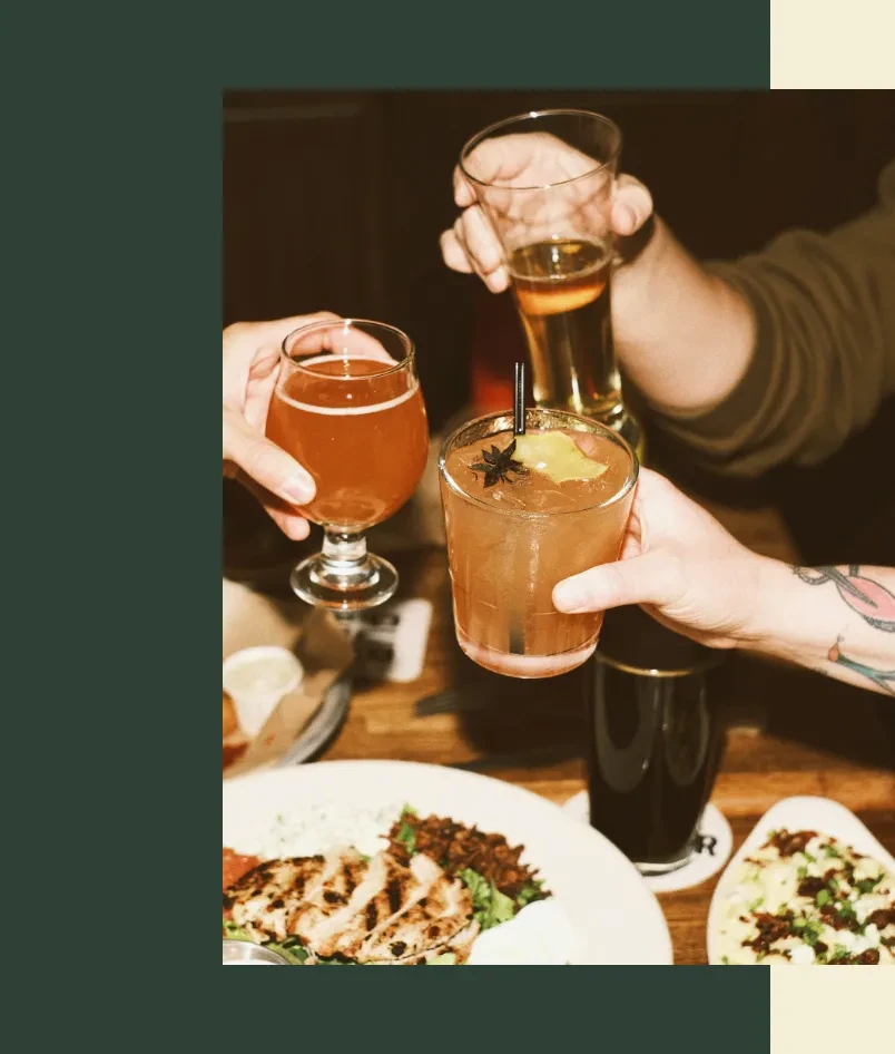 Hands clinking three drinks and food on a cozy restaurant table.