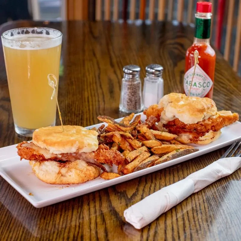 Fried chicken biscuit sandwiches, seasoned fries, beer, and Tabasco on a wooden table.