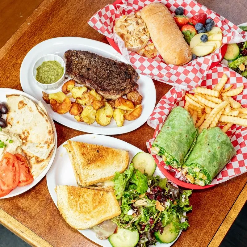 Overhead view of a table with diverse dishes: steak, wraps, sandwich, fruits.