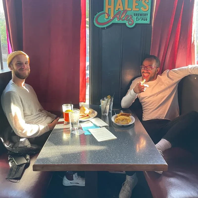 Smiling men in pub booth with drinks, food, Hale's Ales sign.