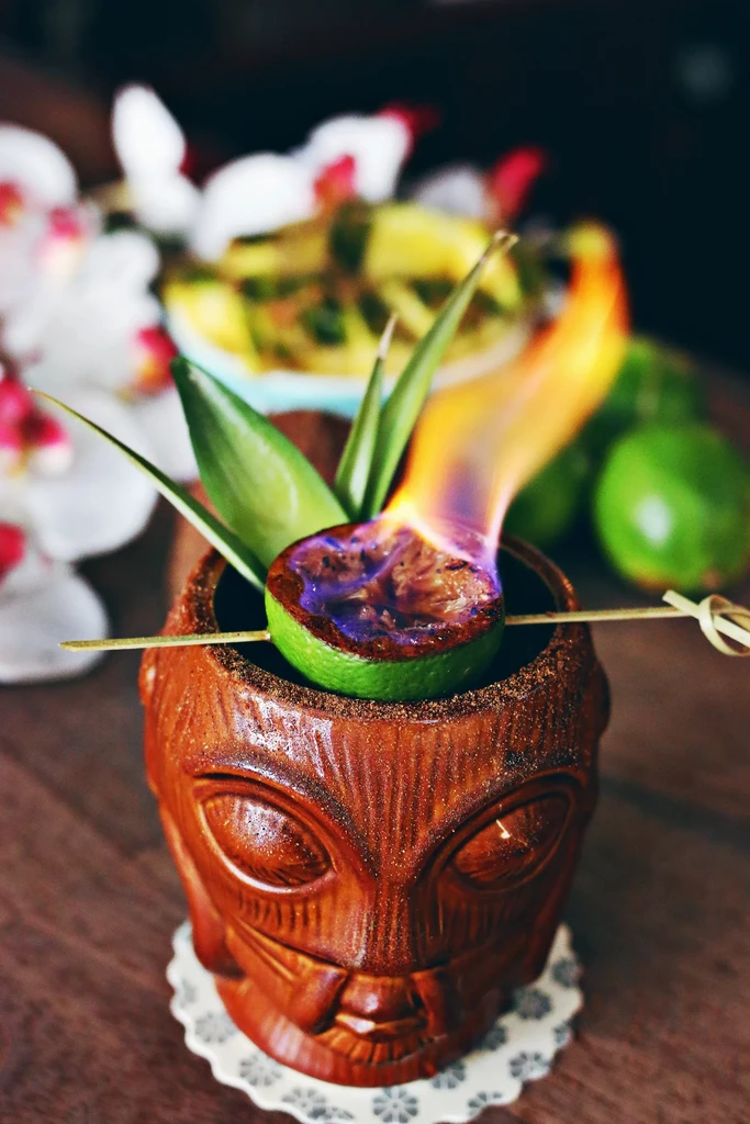 Tiki cocktail in carved mug with burning lime and pineapple leaves.